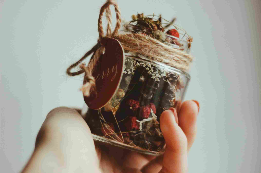 Sebuah stoples kaca berisi potpourri bunga dan rempah kering yang dipegang dengan lembut, sebuah ide DIY yang mudah untuk menciptakan pengalaman aromaterapi di rumah dengan wewangian tahan lama.