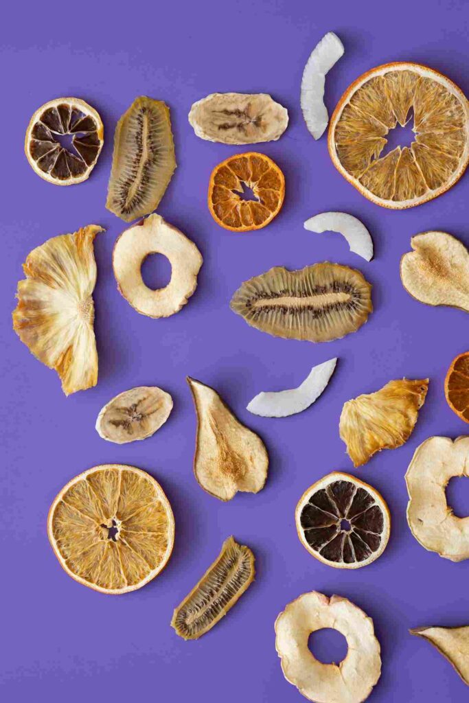 Tampilan atas dari berbagai macam buah kering, termasuk irisan jeruk, kiwi, pisang, nanas, dan kelapa, yang ditata rapi di atas latar belakang ungu cerah, memberikan inspirasi untuk resep trail mix yang penuh warna dan nutrisi.