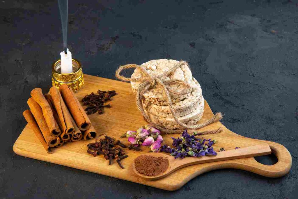 Rangkaian bahan untuk sesi aromaterapi di rumah yang diletakkan di atas talenan kayu, terdiri dari seikat kayu manis, cengkeh, kelopak bunga kering, dan lilin yang menyala, menciptakan suasana relaksasi yang alami.