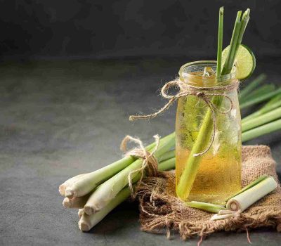 Segelas teh sereh segar dengan madu dan irisan jeruk nipis, dikelilingi oleh batang sereh utuh. Minuman herbal ini menunjukkan salah satu dari banyak manfaat teh sereh untuk kesehatan dan relaksasi tubuh.