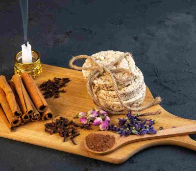 Rangkaian bahan untuk sesi aromaterapi di rumah yang diletakkan di atas talenan kayu, terdiri dari seikat kayu manis, cengkeh, kelopak bunga kering, dan lilin yang menyala, menciptakan suasana relaksasi yang alami.